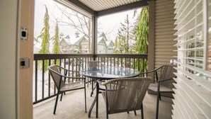 A covered balcony with a glass table and wicker chairs, overlooking a snowy landscape with houses and trees.
