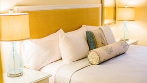 Un lit bien fait, avec du linge de lit blanc et des coussins décoratifs, dont un avec un motif floral. Deux lampes de chevet sont posées sur des tables de chevet blanches.