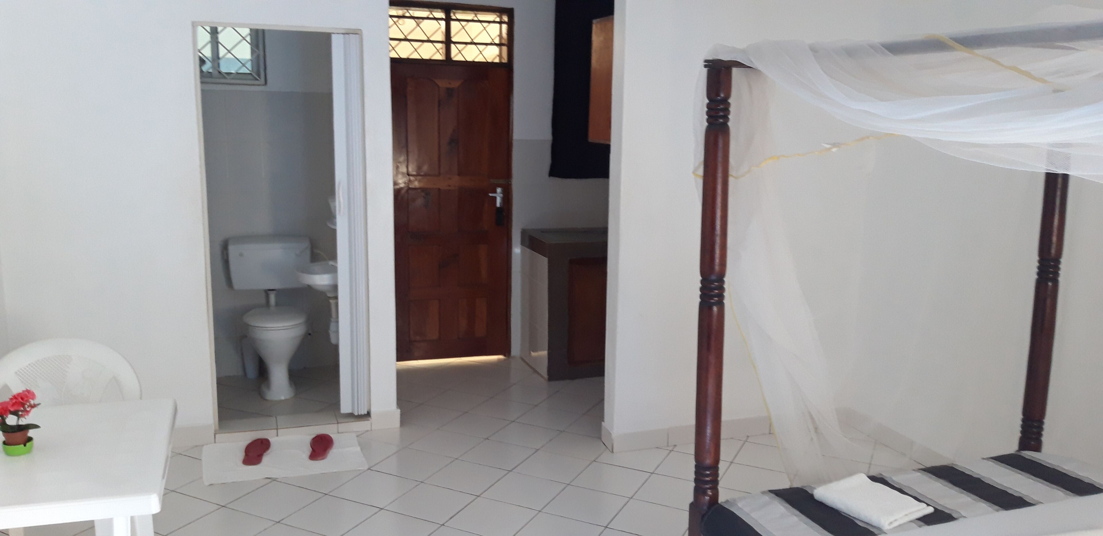 A bedroom with a four-poster bed, a small table with a potted plant, and a view into the bathroom.