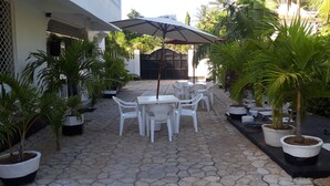 Un patio con suelo de baldosas, sillas de plástico blanco, una mesa y un paraguas grande.