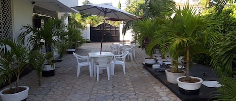 A courtyard with a tiled floor, white plastic chairs, a table, and a large umbrella.