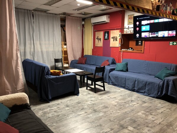 A cozy room with a blue sofa, a small table, and a chair. There is a TV mounted on the wall displaying various channels.