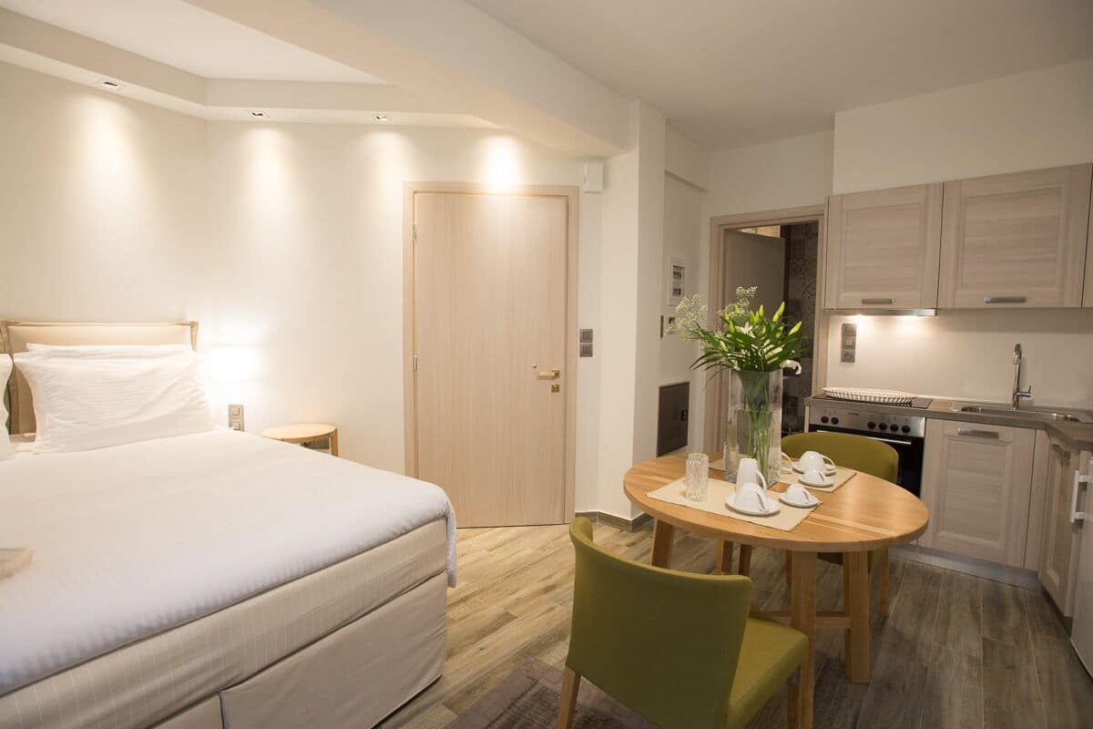 A hotel room with a bed, a dining table with chairs, a kitchen area, and a door.