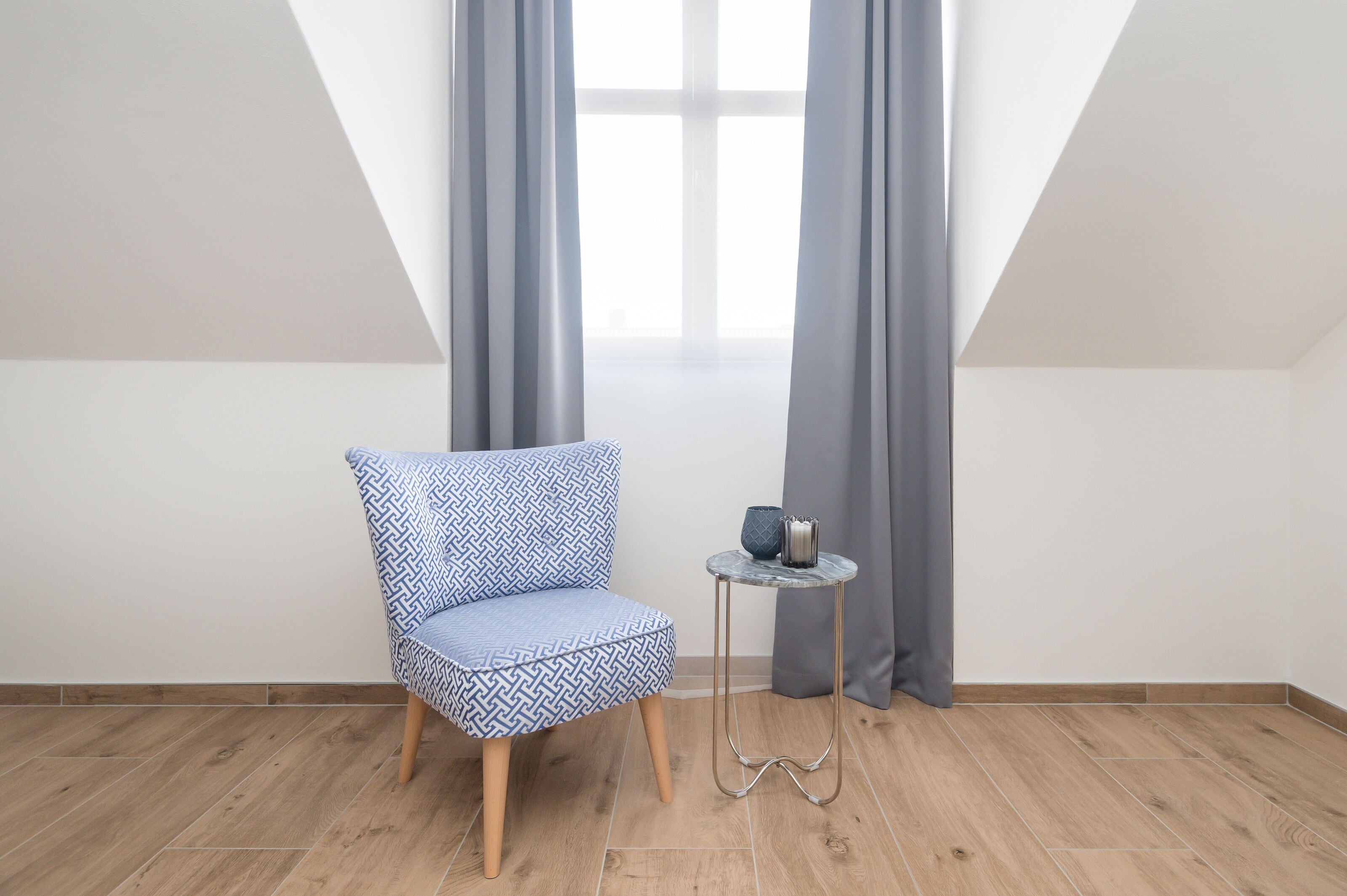 A room with a blue patterned chair, a small round table with a metal base, and grey curtains.