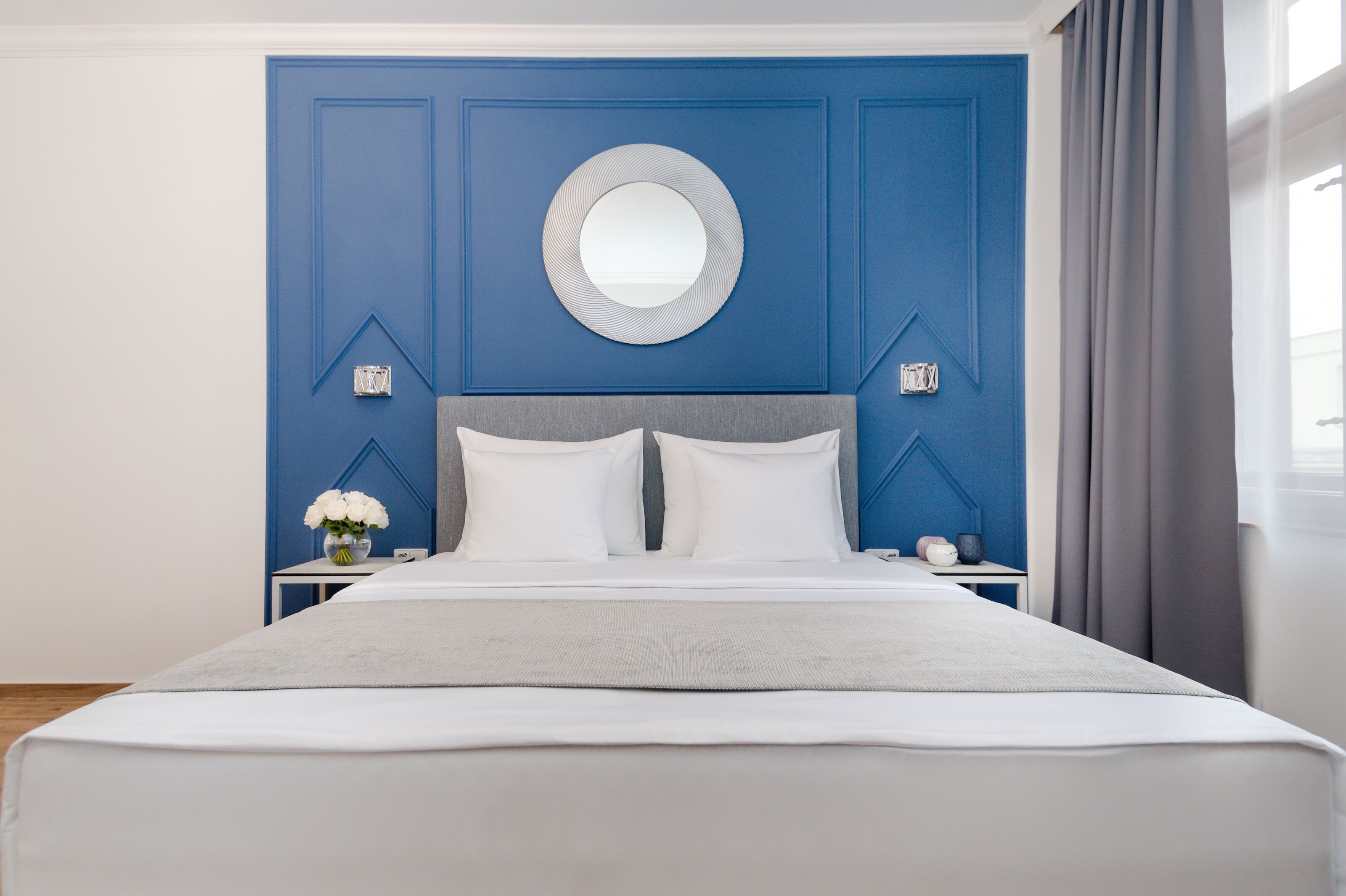 Una cama bien tendida con sábanas blancas, cabecera gris y un espejo redondo en la pared azul sobre ella.