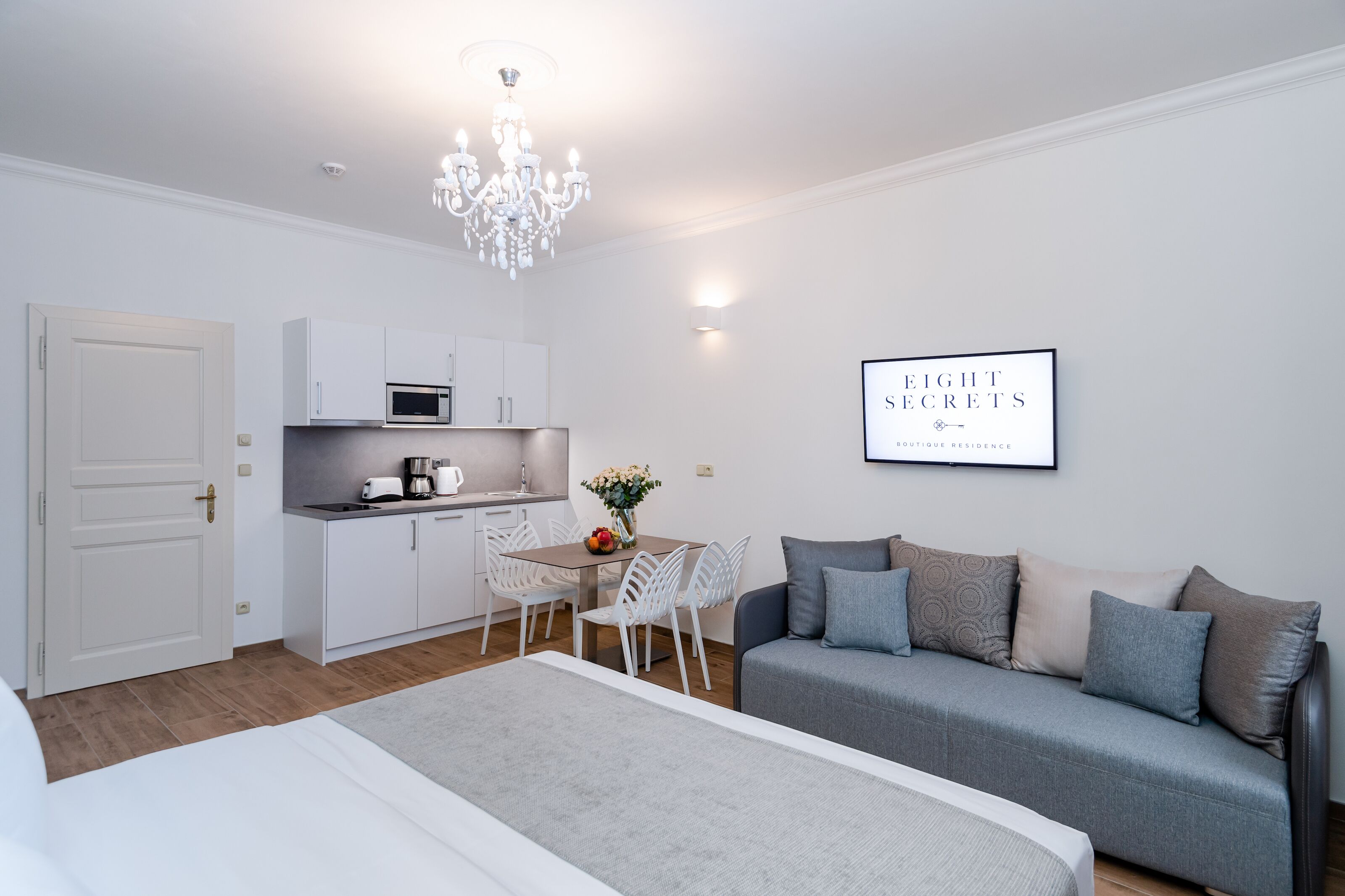 A modern, well-lit room with a kitchenette, a dining area with a table and chairs, a grey sofa, and a bed.