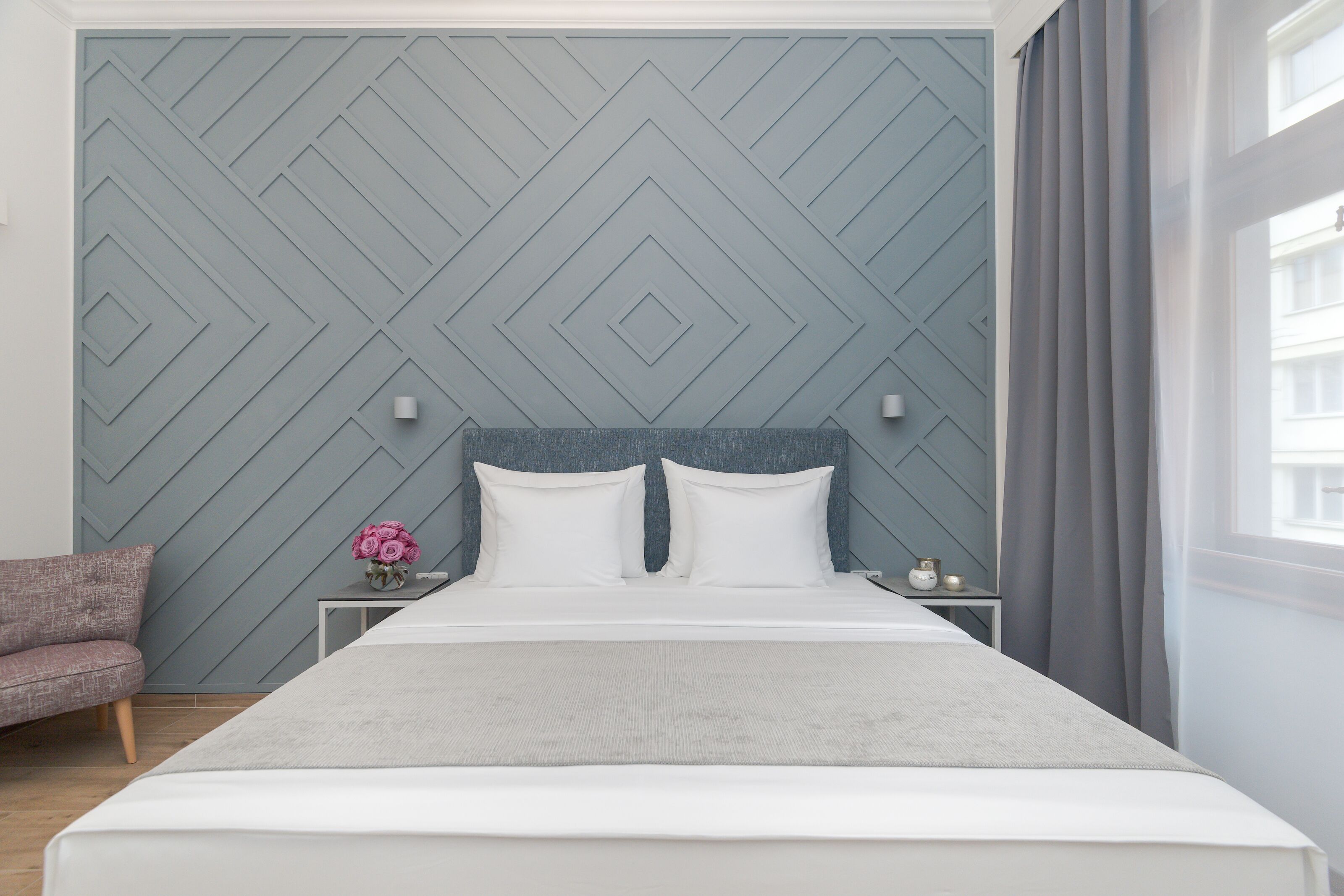 A neatly arranged bedroom with a bed, nightstands, a chair, and a wall with a geometric pattern.