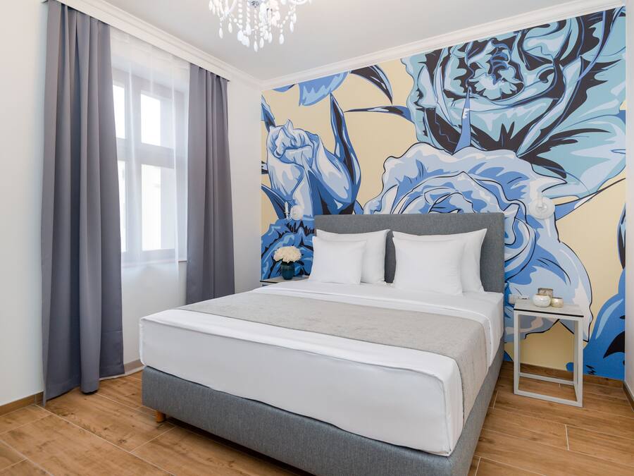Un dormitorio con una cama grande, cortinas grises, un mural floral en la pared y una mesita de noche con un florero.