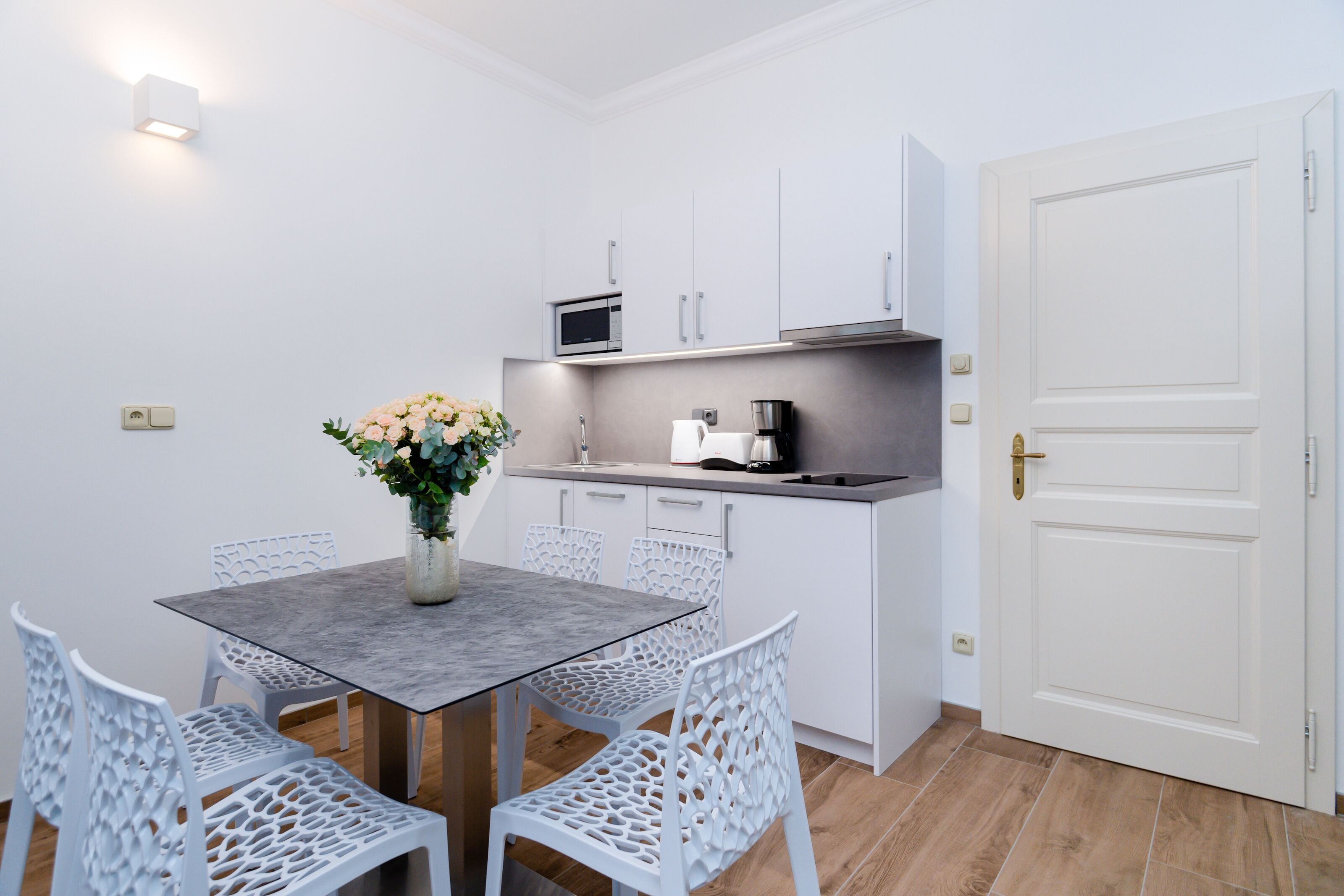 Una cocina compacta con armarios blancos, un microondas y una cafetera. Hay una mesa de comedor con sillas blancas y un jarrón con flores.