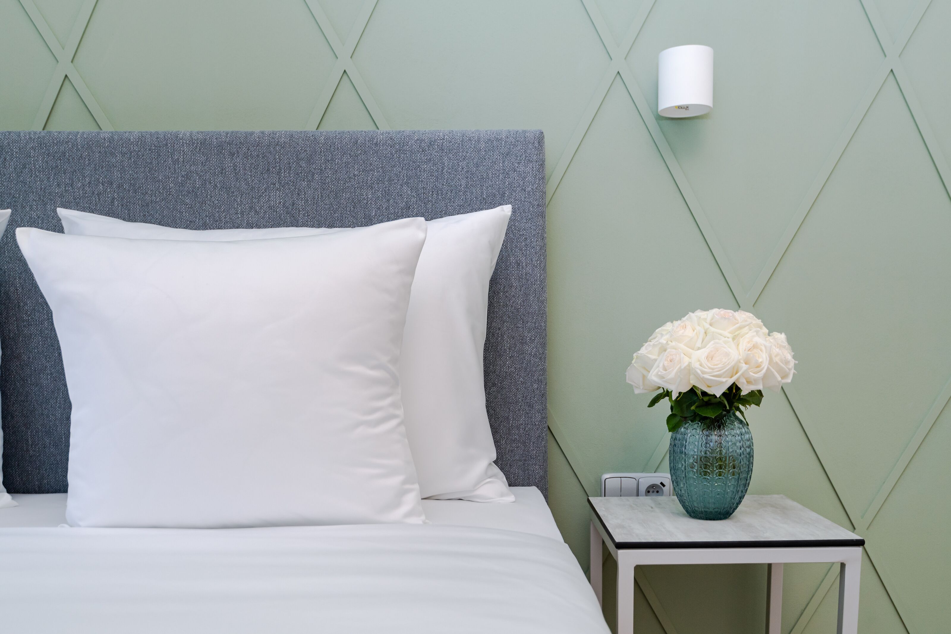 Ein ordentlich bezogenes Bett mit weißen Leinen und Kissen, ein kleiner Tisch mit einer Vase weißer Blumen sowie eine Wandlampe.