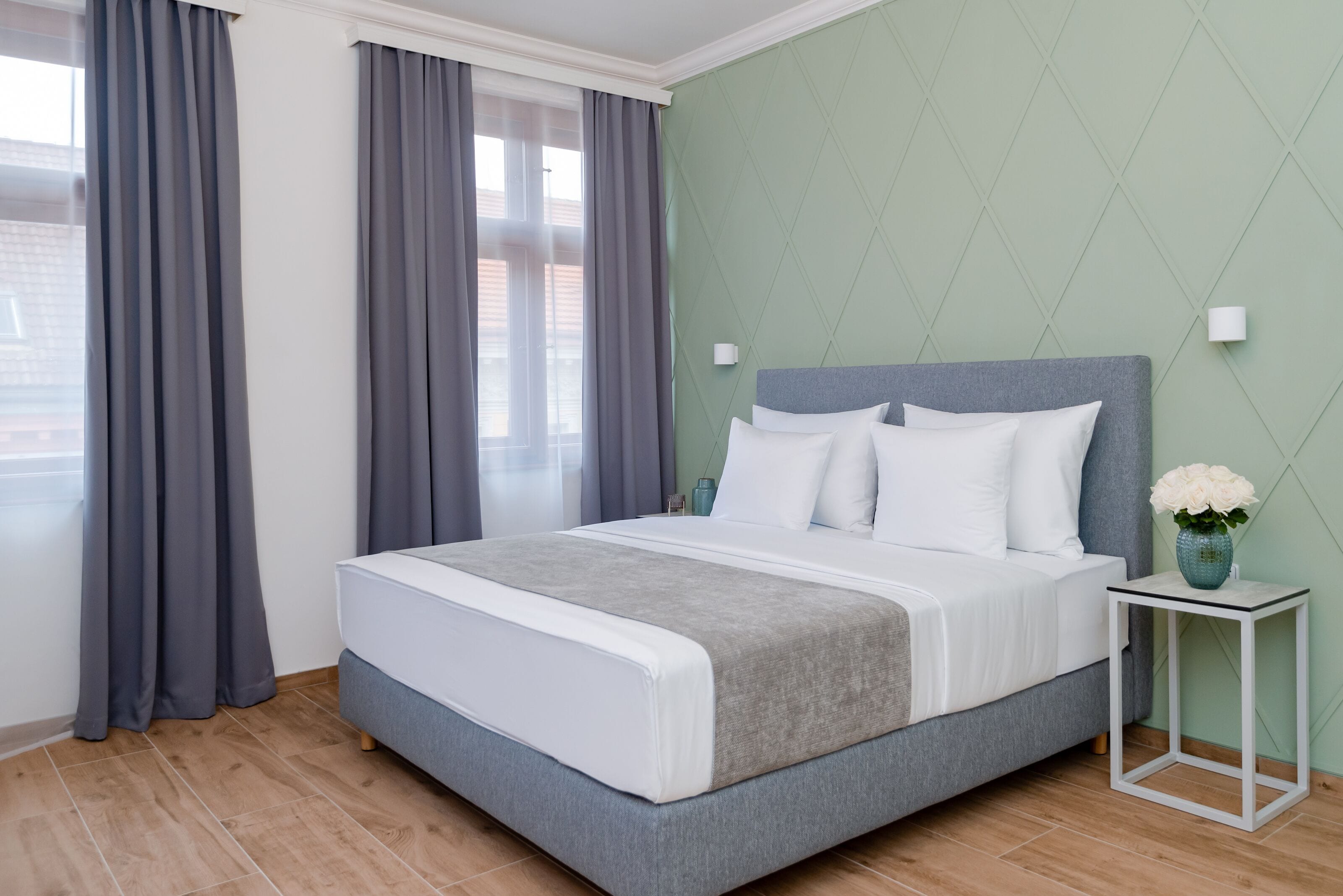 A modern bedroom with a large bed, grey curtains, a green accent wall, and a small bedside table with a vase.