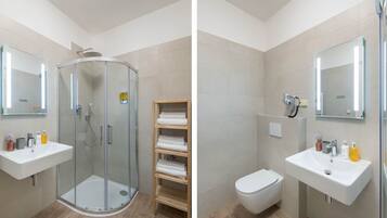 Une salle de bain moderne avec une douche à parois de verre, deux lavabos, des toilettes et une étagère en bois où sont posées des serviettes.