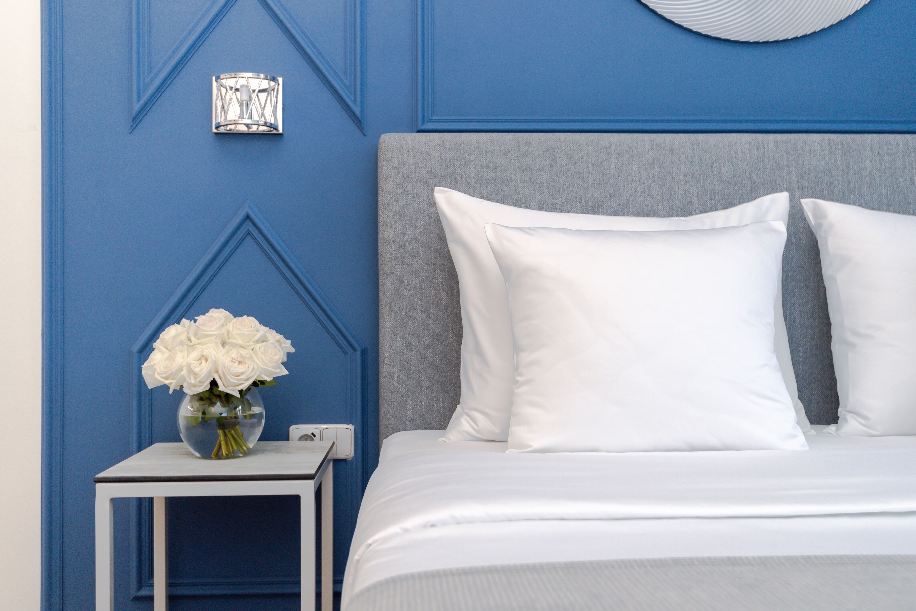 Un lit bien fait, avec des oreillers blancs et une tête de lit grise, une petite table de chevet ornée d’un vase de fleurs blanches, et un mur avec des lambris bleus et un luminaire décoratif.