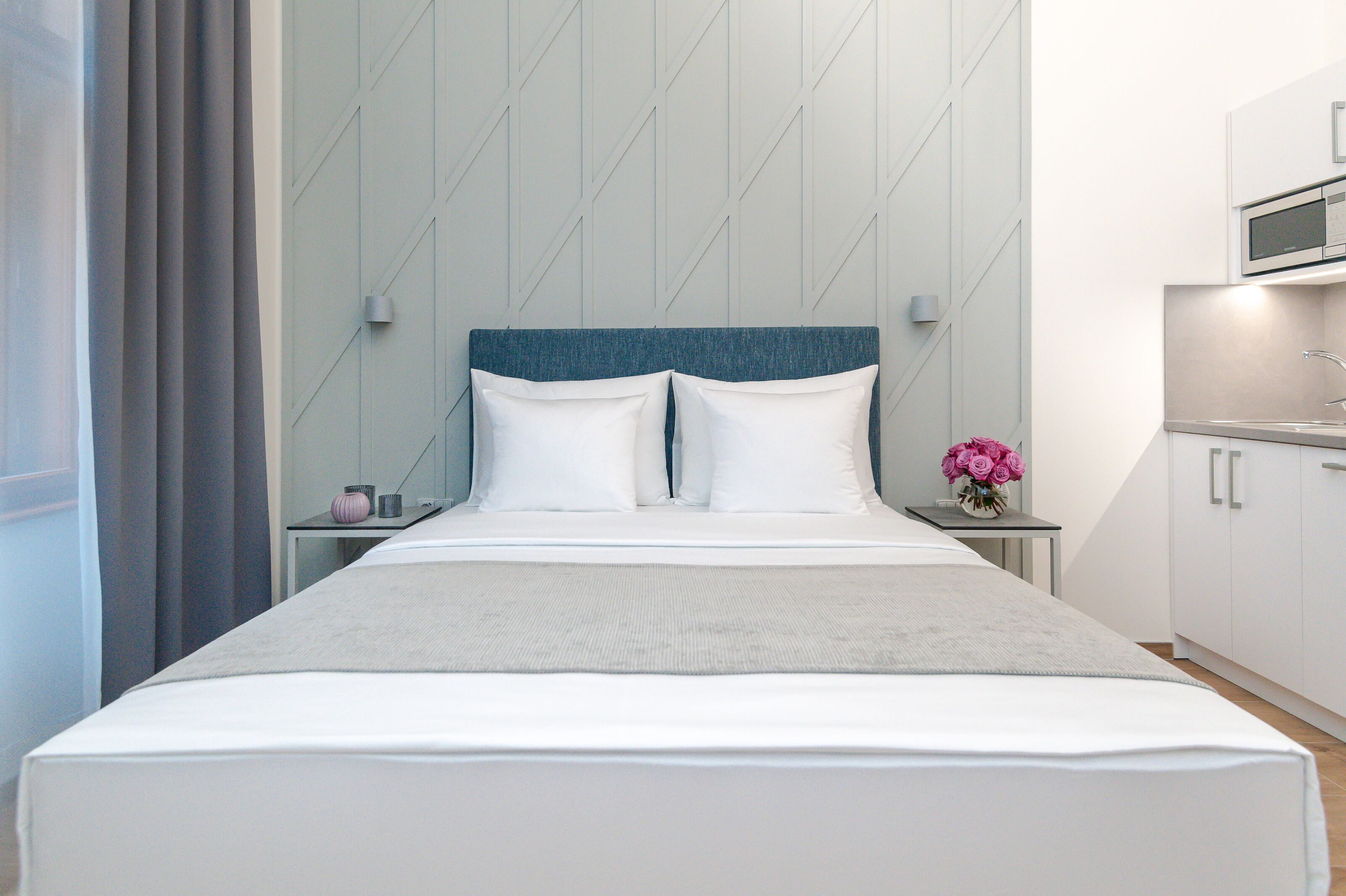 Ein modernes Hotelzimmer mit einem großen Bett, einem kleinen Nachttisch mit Blumenvase und einer Küchenzeile.