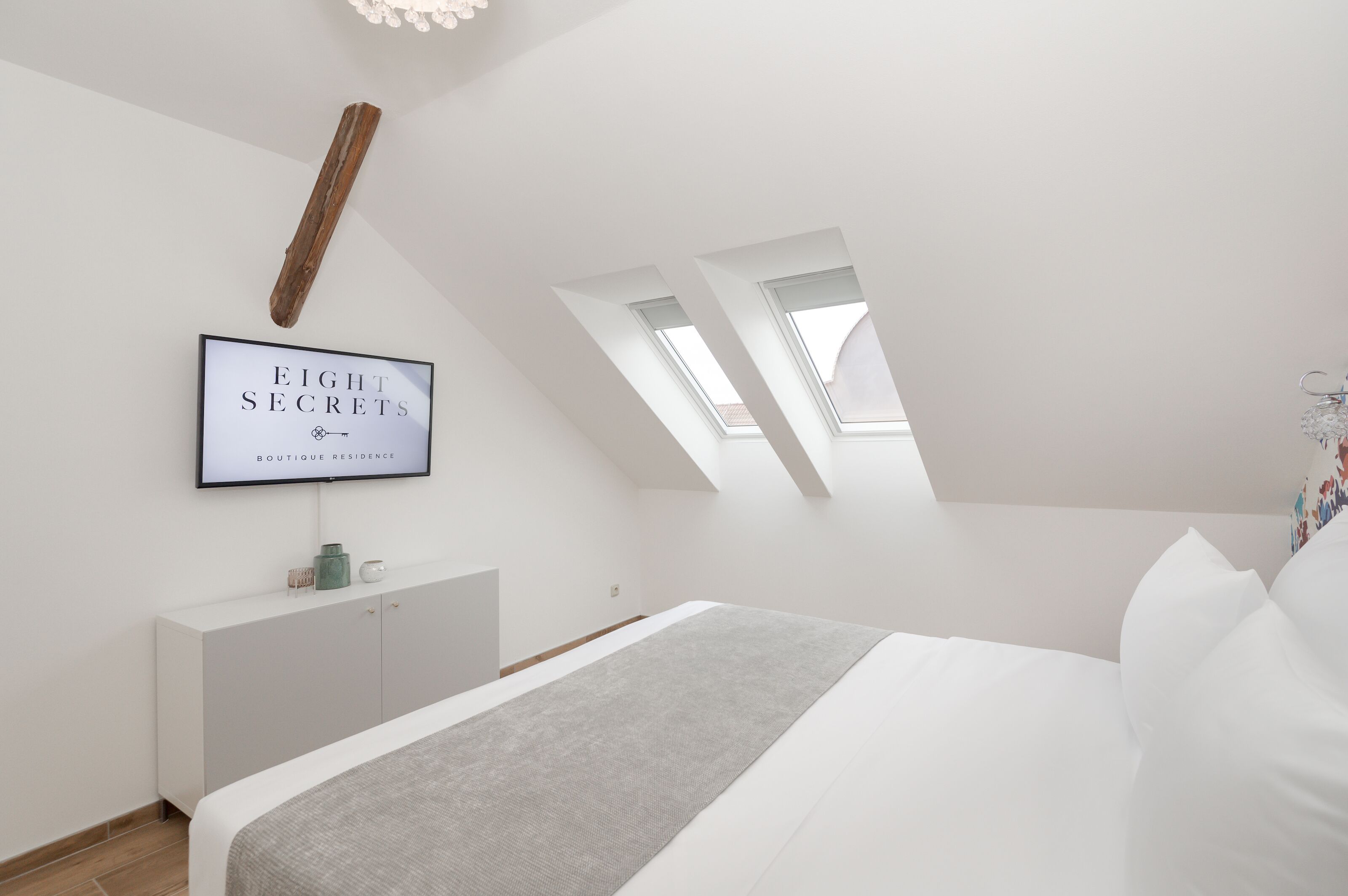 A bedroom with a bed, a white wardrobe, a mounted TV displaying 'EIGHT SECRETS BOUTIQUE RESIDENCE', and skylights.