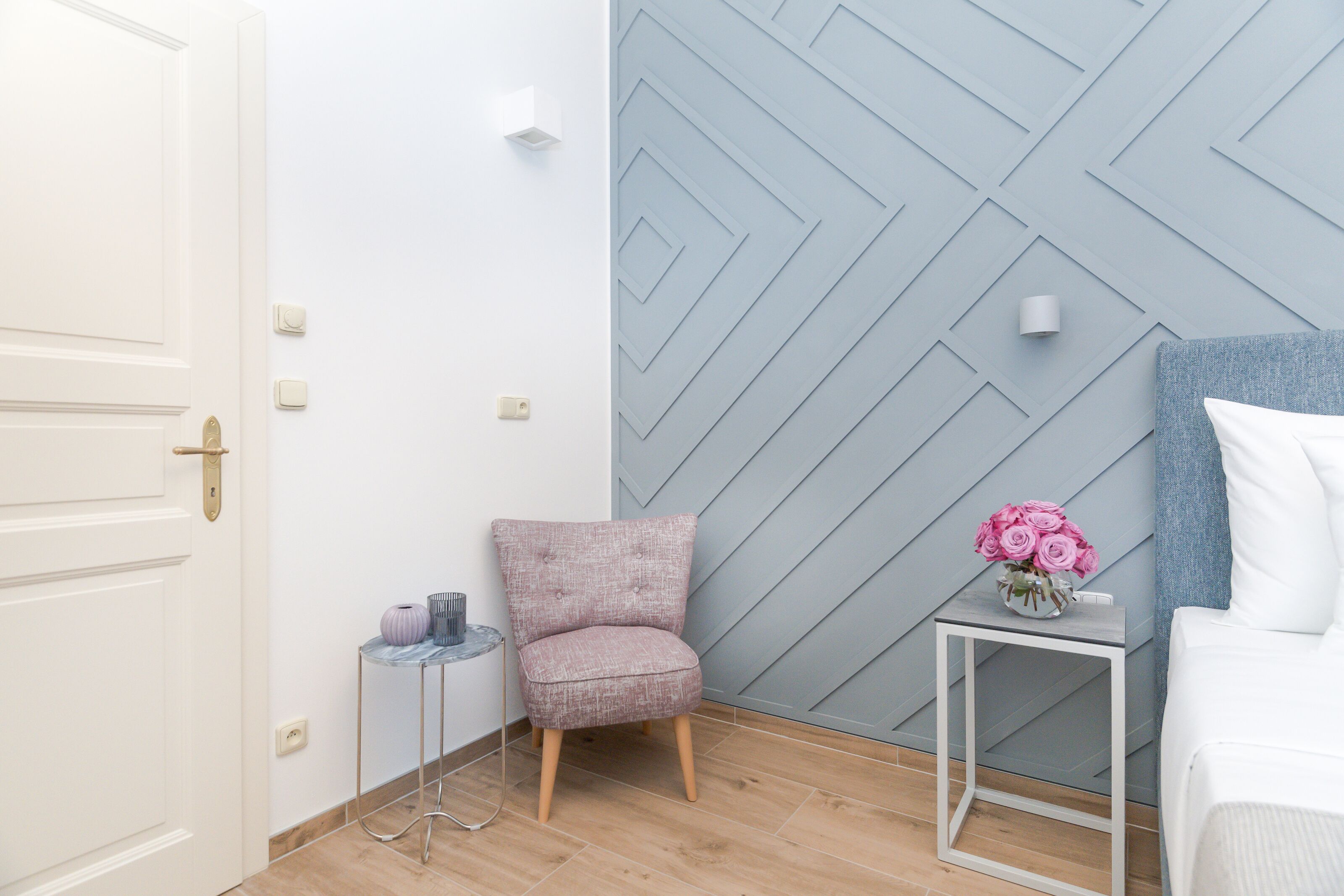 A modern room with a white door, a grey patterned wall, a small side table with a vase of pink flowers, and a white bed with a blue headboard.