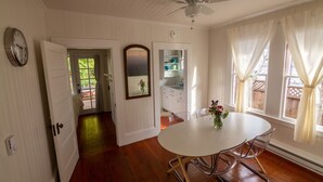 Un coin repas avec une table et des chaises blanches, un ventilateur de plafond et une vue sur le jardin à travers de grandes fenêtres.