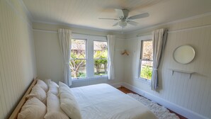 Une chambre à coucher avec un lit, un ventilateur de plafond, deux fenêtres avec des rideaux, un tapis blanc et un miroir fixé au mur.