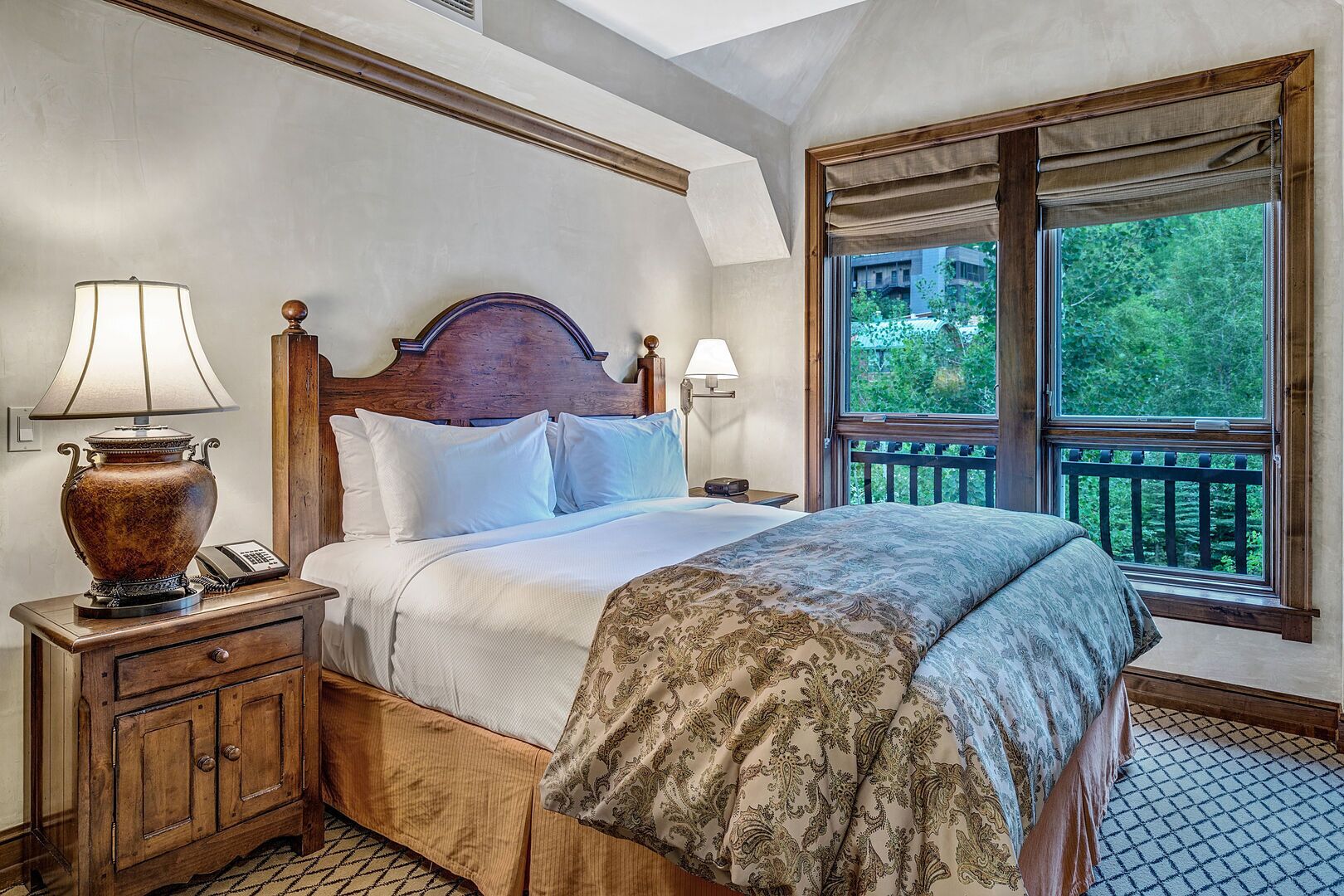 A bedroom with a large bed, two bedside lamps, a wooden nightstand, a telephone, and a window with a view of greenery.