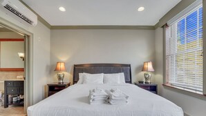 Una cama bien hecha con sábanas y toallas blancas, un cabecero, dos lámparas de noche y una ventana con persianas.