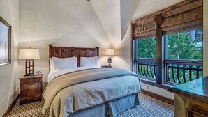 A bedroom with a large bed, two bedside lamps, a nightstand, a window with a view of greenery, and a wooden headboard.