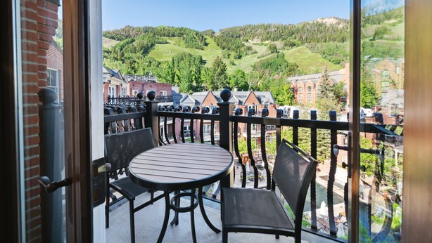 A balcony with a round table and chairs, offering a view of a green valley and buildings.