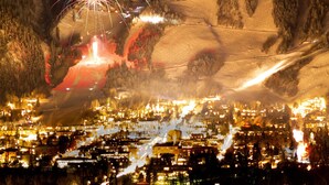 Lumipeitteisen vuoristokaupungin näkymä yöllä, taivaalla ilotulitus ja alapuolella valaistu kaupunki.