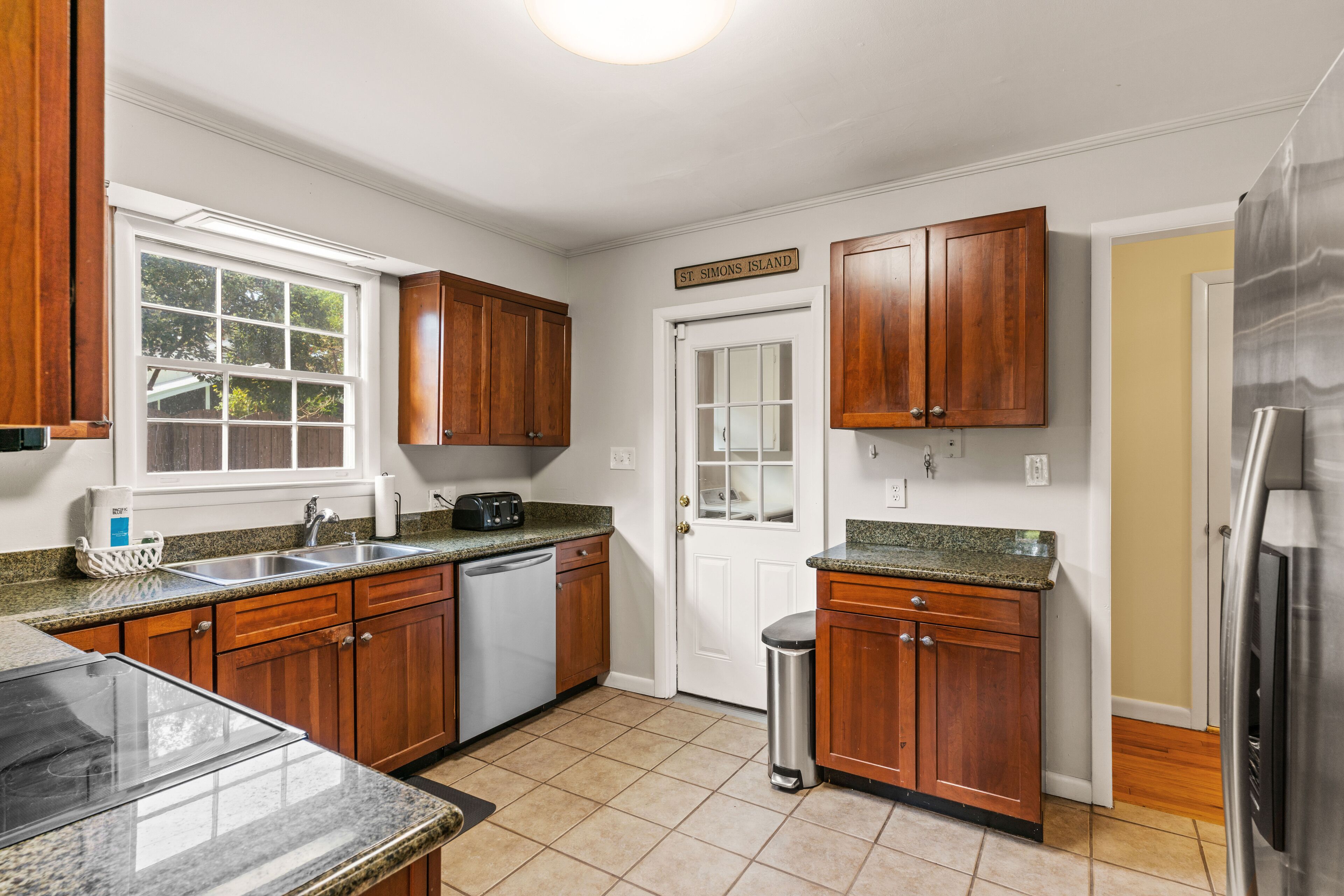 A kitchen with wooden cabinets, a stainless steel refrigerator, and a window with a view of trees.