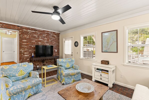A living room with a brick wall, a ceiling fan, a TV, two patterned armchairs, a coffee table, and a white cabinet.