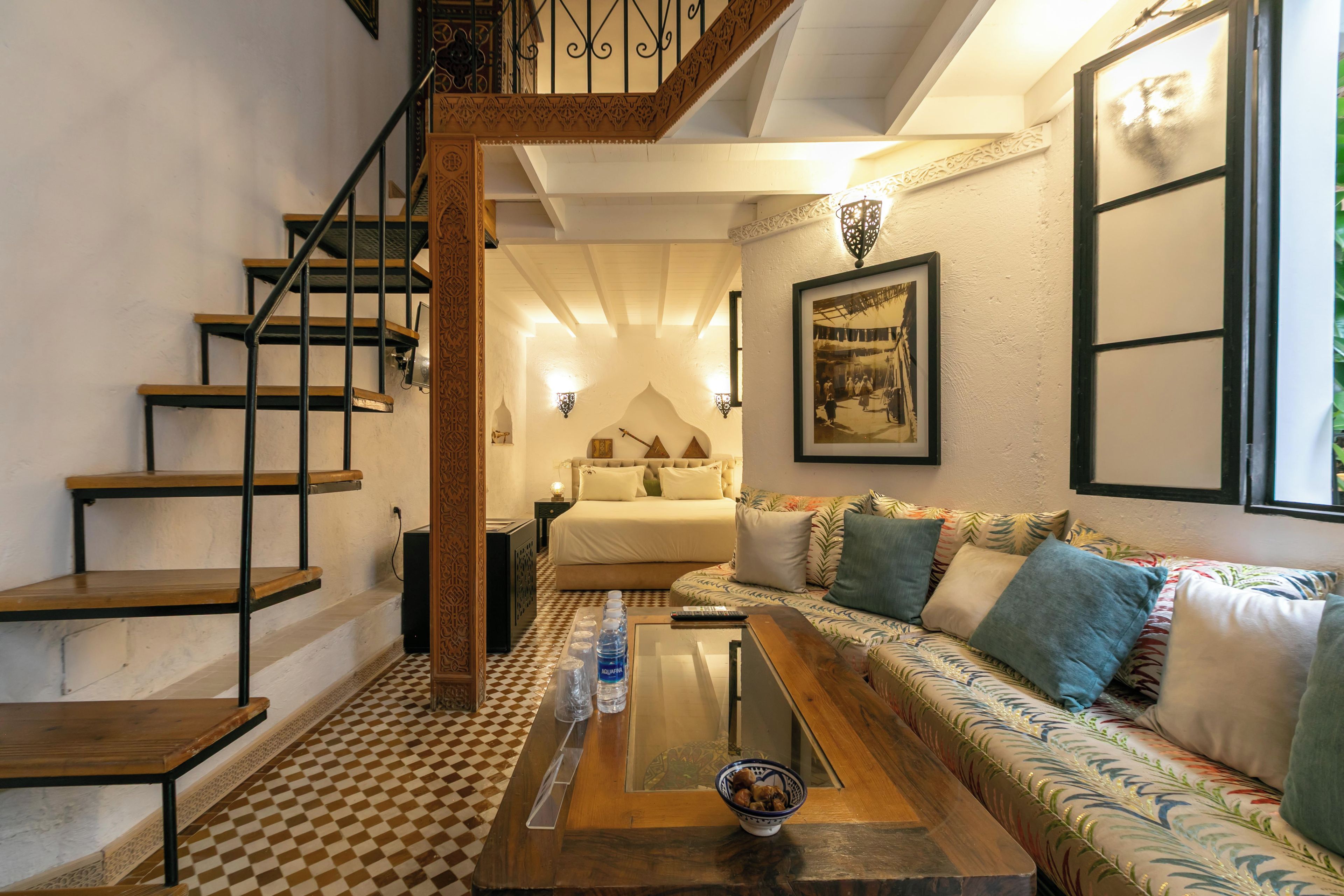 A hotel room with a checkered floor, a sofa with patterned cushions, a bed with a headboard, a staircase with wooden steps, and a wall-mounted picture.