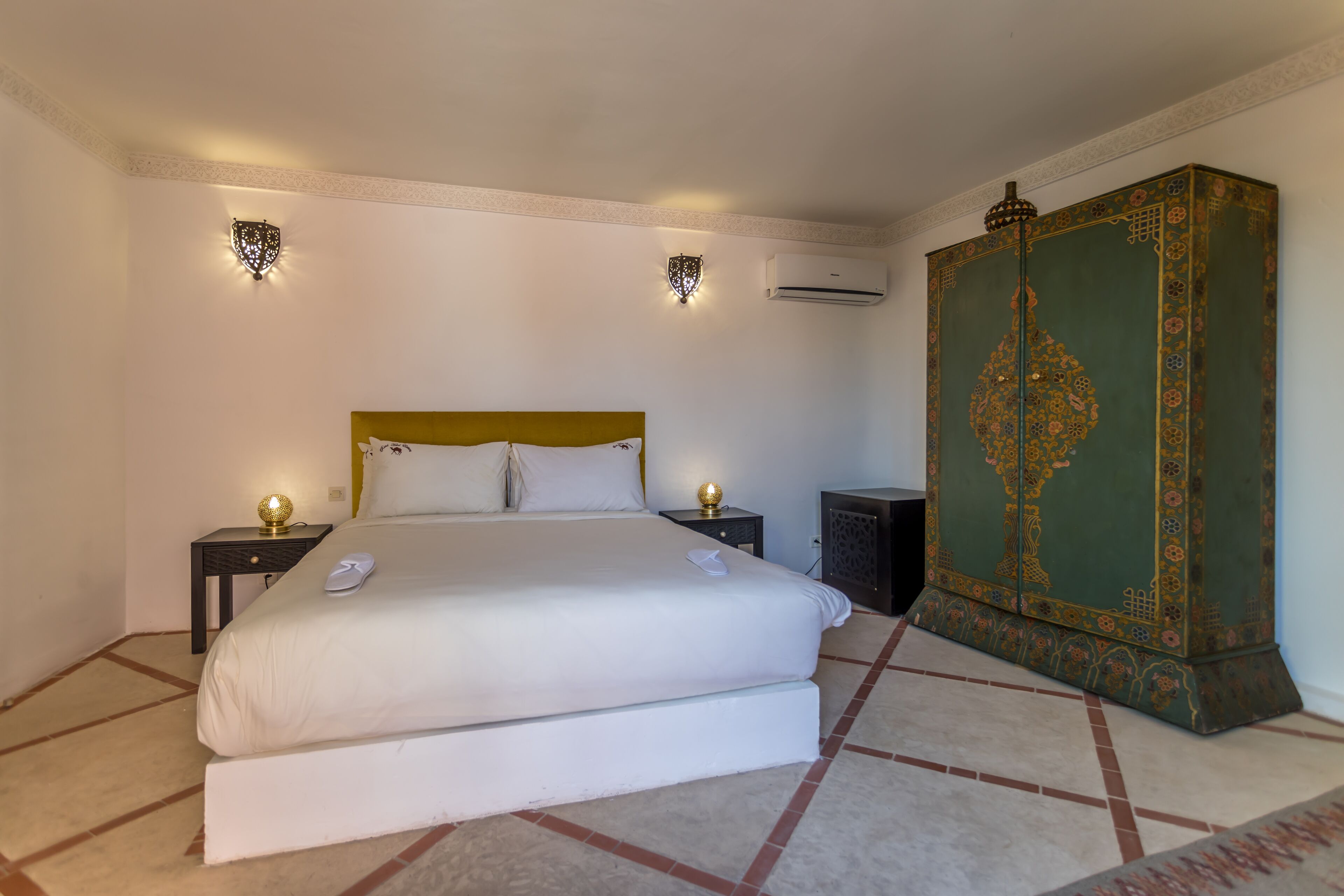 A hotel room with a large bed, two bedside tables with lamps, an air conditioning unit, and a decorative cabinet.