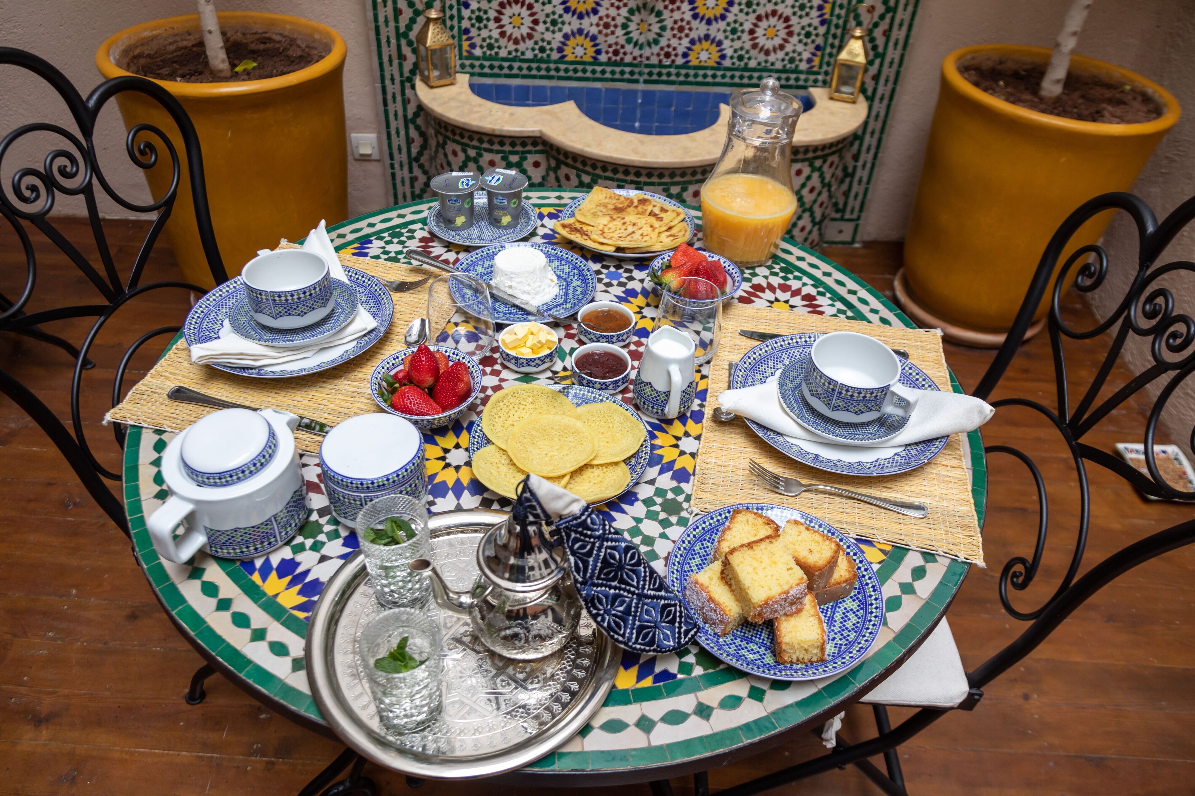 A round table with a Moroccan-style tablecloth, surrounded by chairs, and filled with various breakfast items including bread, fruit, and beverages.