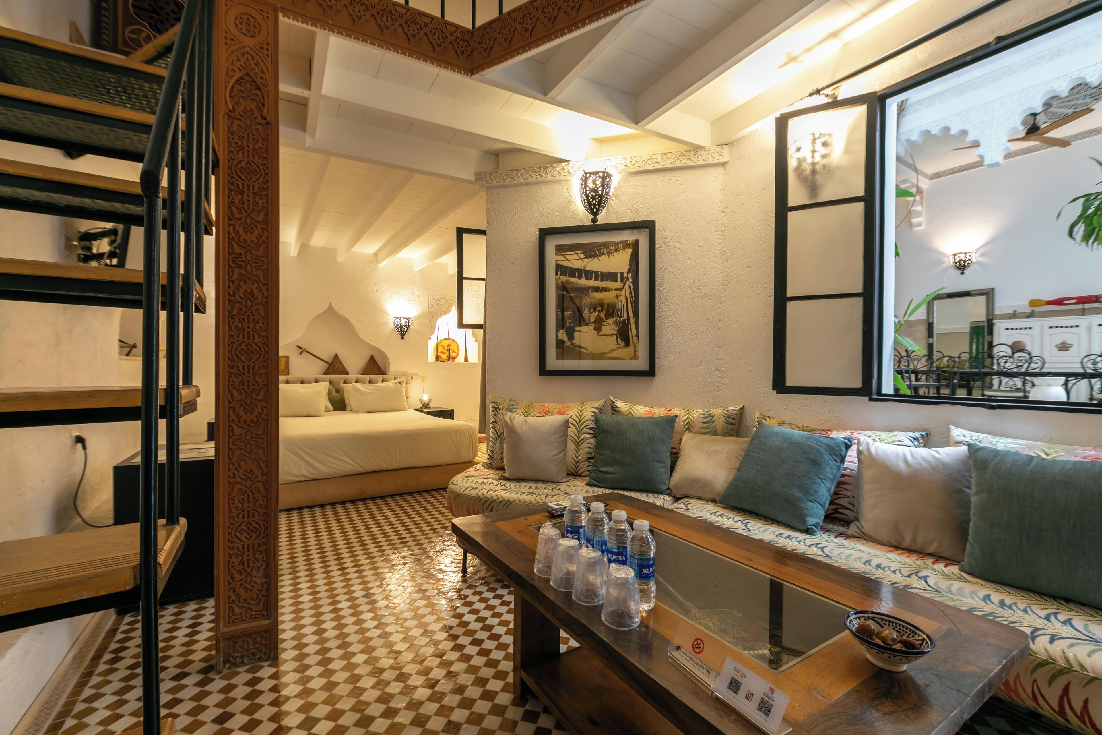 Una habitación de hotel con piso a cuadros, una cama con sábanas blancas, un sofá con cojines estampados, una mesa de centro con botellas de agua y un cuadro en la pared.