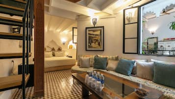 Una habitación de hotel con piso a cuadros, una cama con sábanas blancas, un sofá con cojines estampados, una mesa de centro con botellas de agua y un cuadro en la pared.