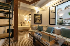 Une chambre d’hôtel avec un sol à carreaux, un lit avec du linge de maison blanc, un canapé avec des coussins à motifs, une table basse avec des bouteilles d’eau et un tableau encadré au mur.