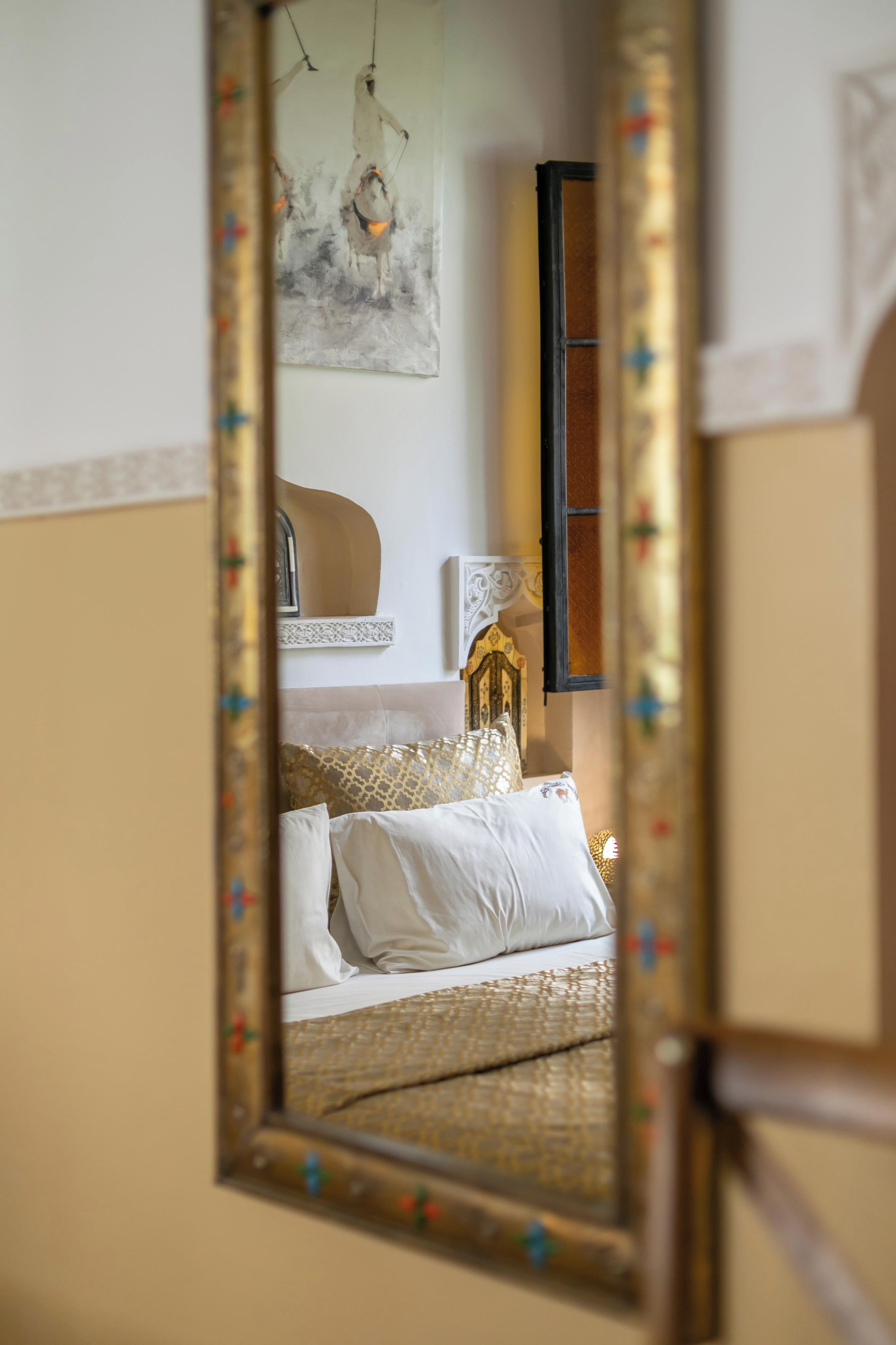 Un dormitorio con cama, almohadas, mesita de noche, espejo y una obra de arte colgada en la pared.