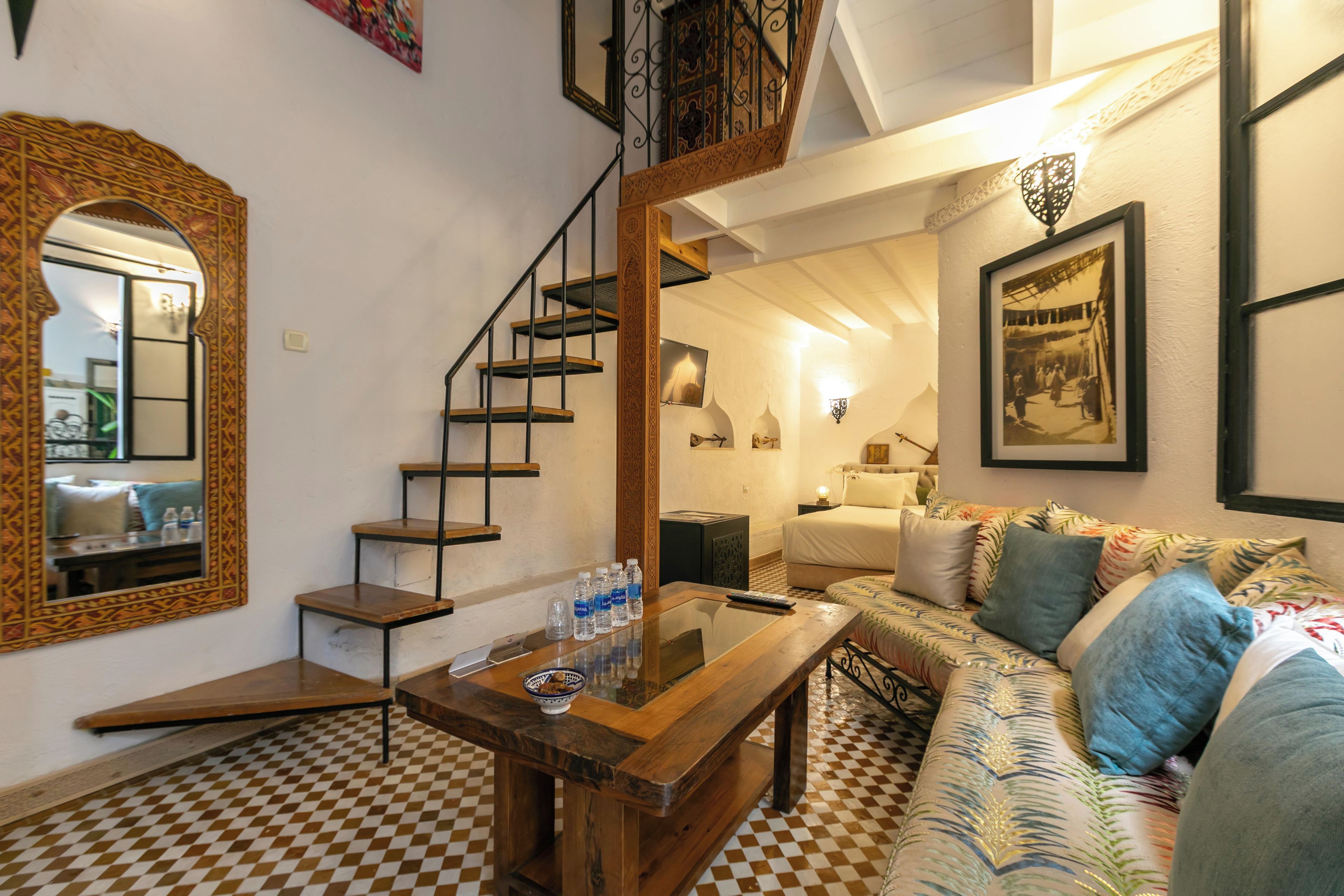 A room with a checkered floor, a wooden table with water bottles, a sofa with patterned cushions, a staircase with a railing, and a mirror with a decorative frame.