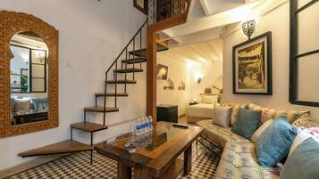 A room with a checkered floor, a wooden table with water bottles, a sofa with patterned cushions, a staircase with a railing, and a mirror with a decorative frame.
