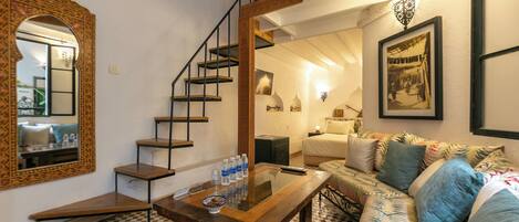 A room with a checkered floor, a wooden table with water bottles, a sofa with patterned cushions, a staircase with a railing, and a mirror with a decorative frame.