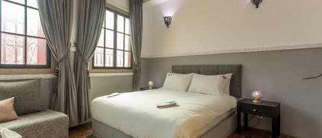 A neatly arranged bedroom with a bed, bedside tables, a sofa, and a window with curtains.