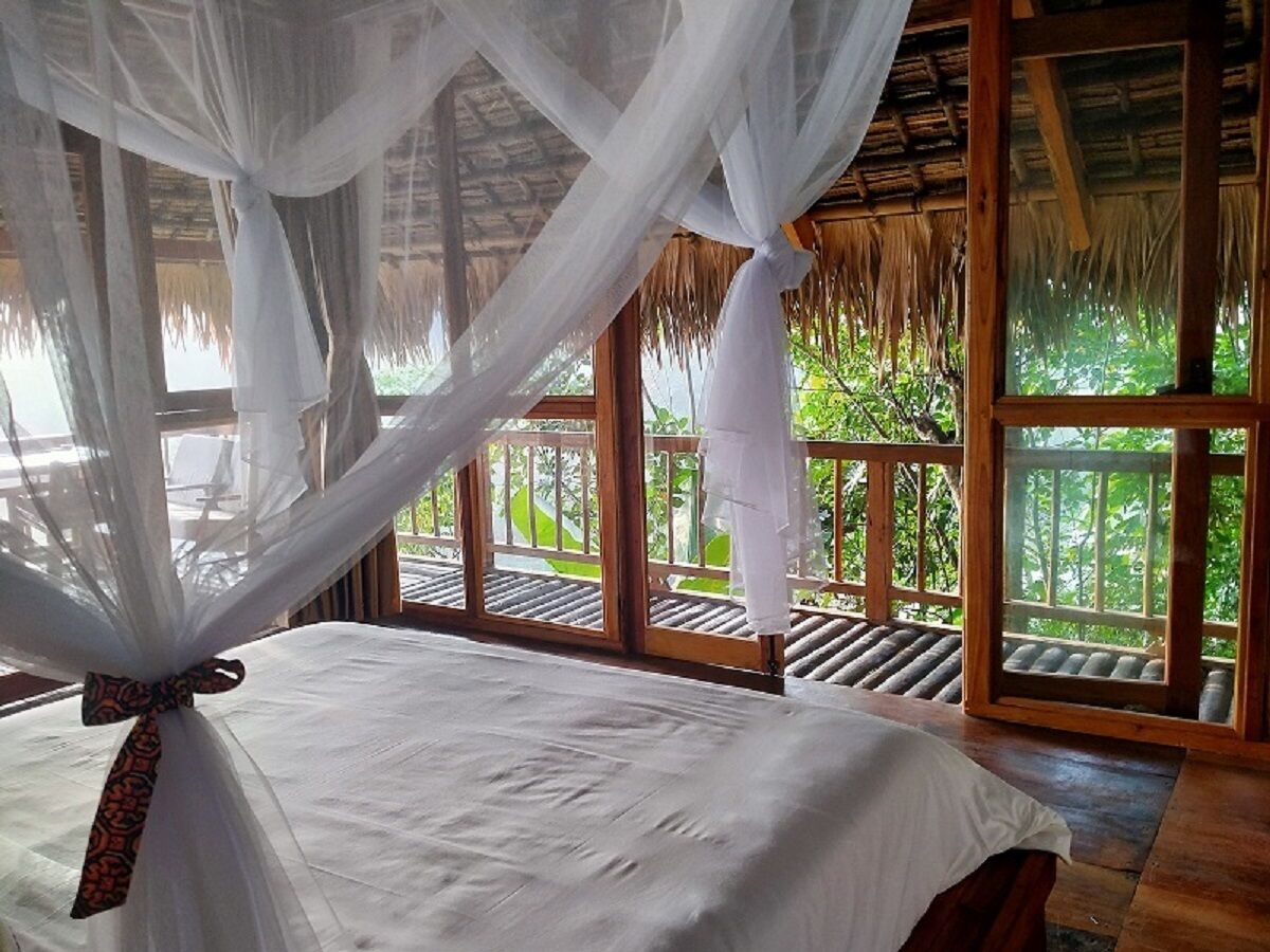 A bedroom with a canopy bed, wooden flooring, and large windows with a view of greenery.