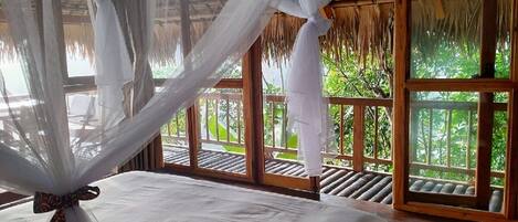 A bedroom with a canopy bed, wooden flooring, and large windows with a view of greenery.