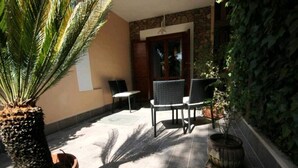 En udendørs patio med stenvæg, to fletstole, en plantekasse og flisegulv.