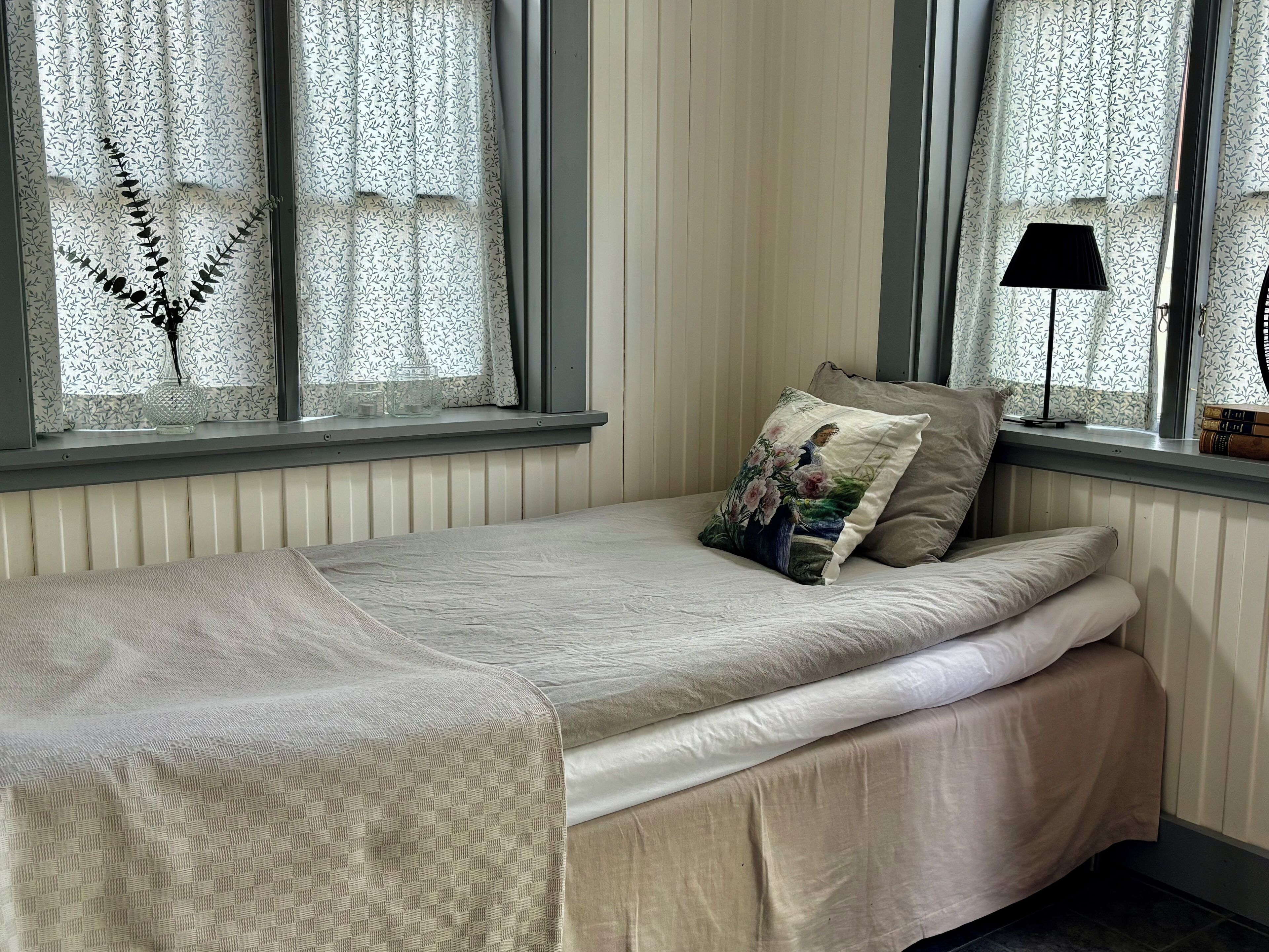A single bed with a patterned bedspread, a decorative pillow with a floral design, and a window with sheer curtains and a fern decoration.