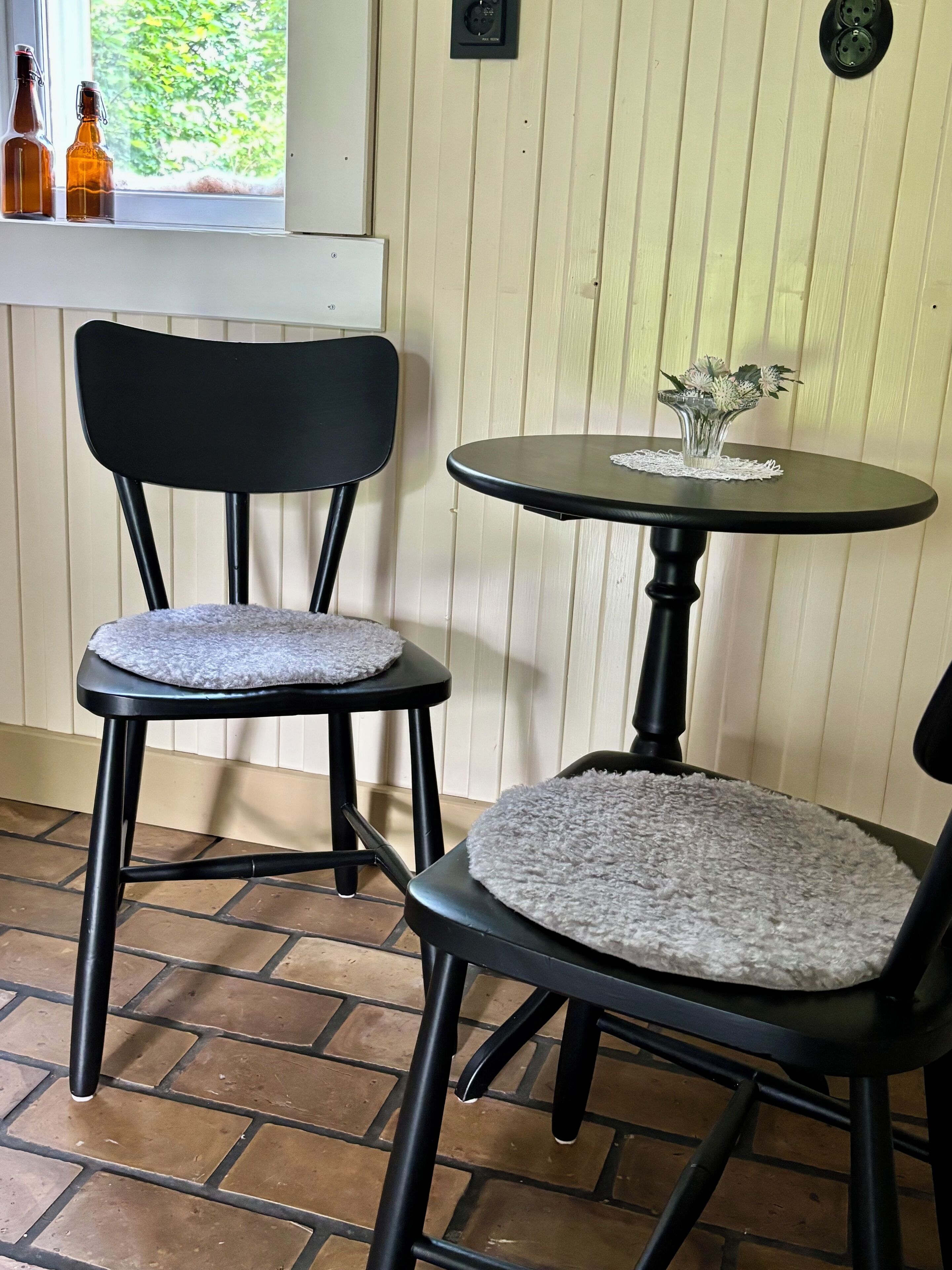 Deux chaises noires avec des coussins ronds blancs, une petite table ronde surmontée d’un chemin de table en dentelle et une fenêtre dont l’appui est orné de bouteilles.