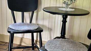 Deux chaises noires avec des coussins ronds blancs, une petite table ronde surmontée d’un chemin de table en dentelle et une fenêtre dont l’appui est orné de bouteilles.