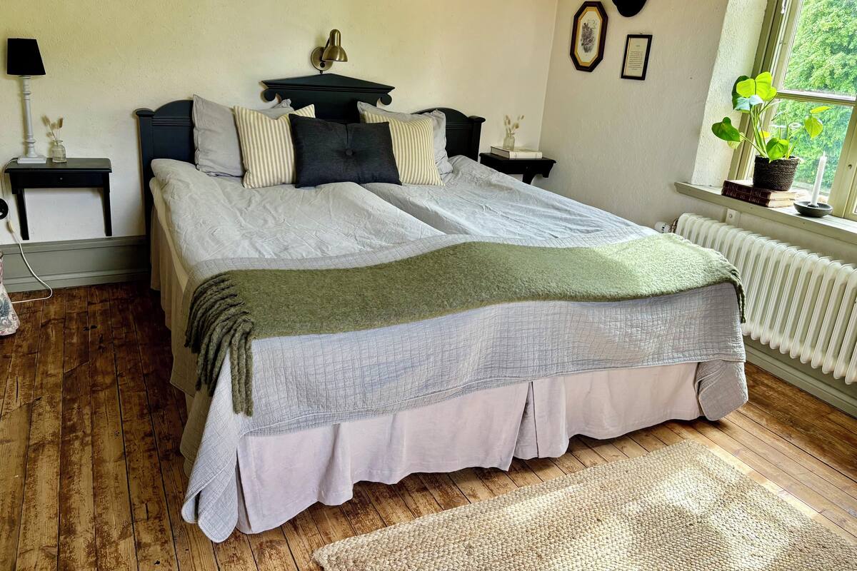 A bedroom with a large bed, two bedside tables, a radiator, and a window with a view of greenery.