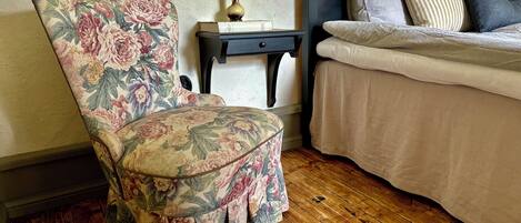 A bedroom with a floral-patterned armchair, a bedside table with a vase, and a bed with pillows.