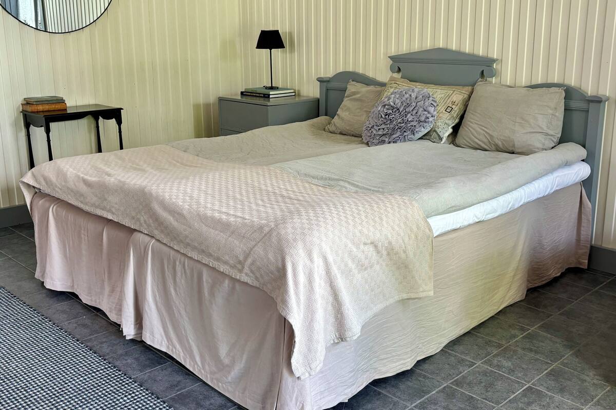 A neatly made bed with a beige bedspread and pillows, a bedside table with a lamp, and a small stack of books on the wall.