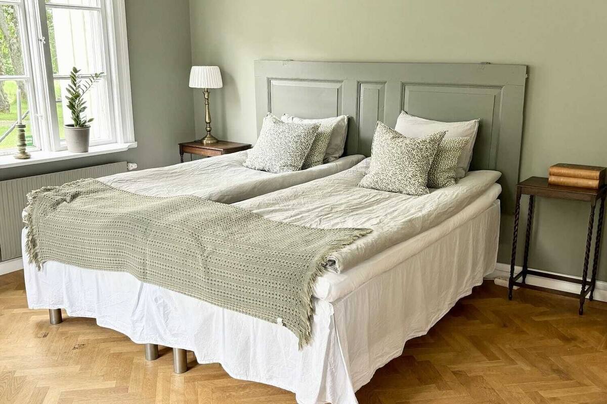 A double bed with a white and green blanket, two bedside tables with lamps, and a window with a plant.